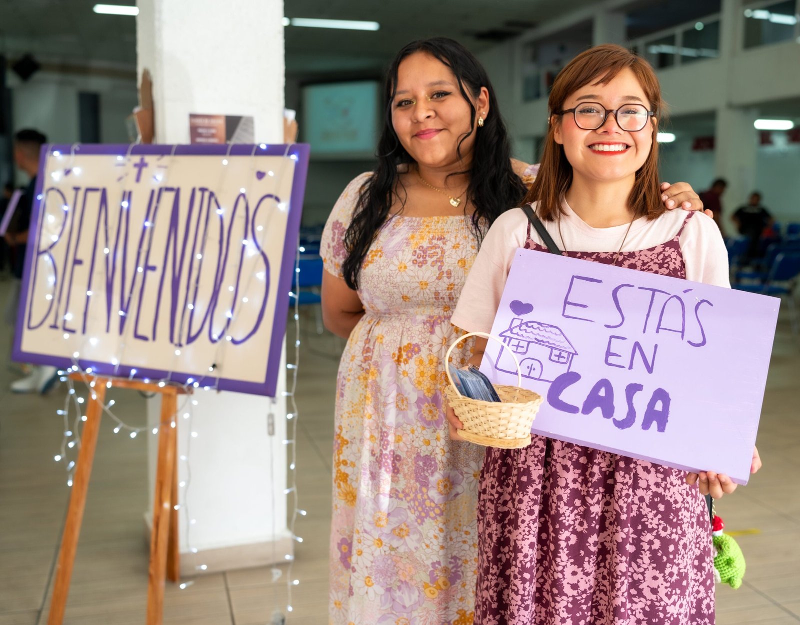 Two women warmly greet visitors at a community event in Ciudad de México with signs saying 'Bienvenidos' and 'Estás en Casa'.